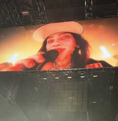 a woman in a white hat is on a large screen with microphones and lights behind her