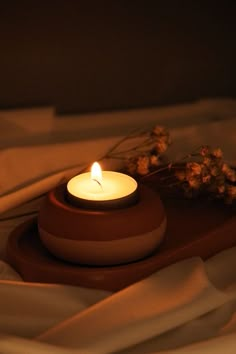 a lit candle sitting on top of a wooden tray next to dried flowers and white sheets