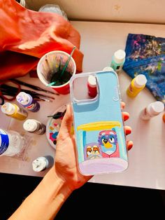 a person holding up a phone case with an owl painting on it and other art supplies