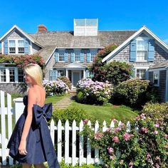 a woman walking past a white picket fence in front of a house with pink flowers