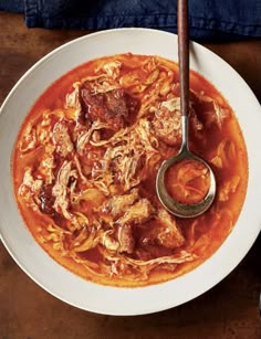 a white bowl filled with meat and tomato soup on top of a wooden table next to a spoon