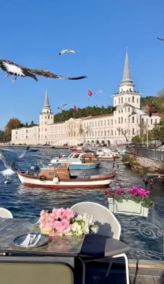 birds are flying over the water and boats on the river in front of a large white building