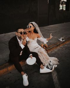 a bride and groom are sitting on the curb drinking from wine glasses while holding plates