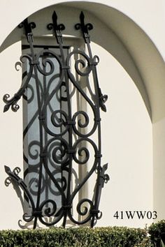 an arched window with wrought iron work on the side of a white wall and green shrubbery