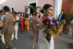 a woman in a white dress holding a bouquet and walking down the street with other people