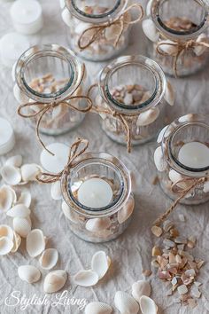 small glass jars filled with seashells and candles on a white cloth covered table