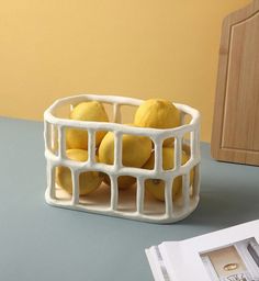 three lemons in a white basket sitting on a table next to an open magazine