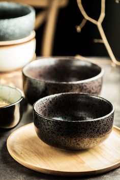 three black bowls sitting on top of a wooden plate next to other bowls and saucers