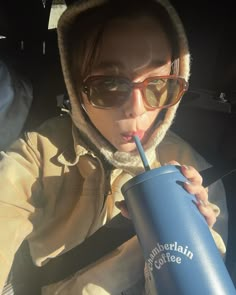 a woman in sunglasses drinking from a blue coffee cup while sitting in the back seat of a car