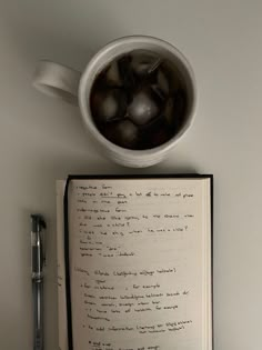 an open book sitting on top of a table next to a pen and cup filled with liquid