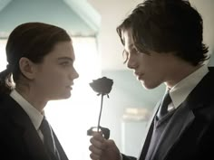 two people in suits are facing each other and one person is holding a rose up to his ear