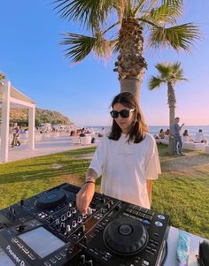 a woman standing next to a dj set up in front of a palm tree on the beach