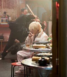 a woman sitting at a table in front of a man with a baseball bat over her head