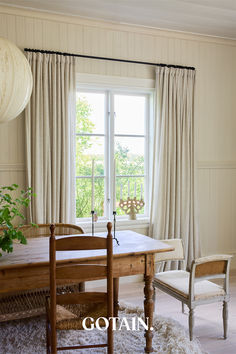 a dining room table and chairs in front of a window with white drapes on the windowsill