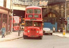 a red double decker bus driving down a street next to tall buildings and people walking on the sidewalk