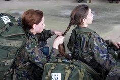 two women in uniform sitting next to each other with backpacks on their backs and one woman tying her hair