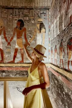 a woman in a yellow dress and straw hat standing next to an egyptian wall painting