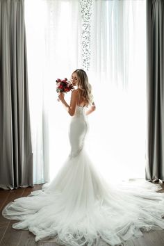 a woman in a white wedding dress standing next to a window with sheer drapes