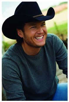 a man wearing a black cowboy hat sitting on a wooden bench smiling at the camera