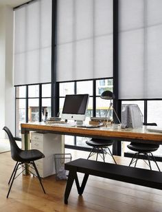 a desk with two computers on it in front of a window covered by roller shades