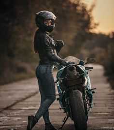 a woman standing next to a motorcycle on a road with trees in the background at sunset