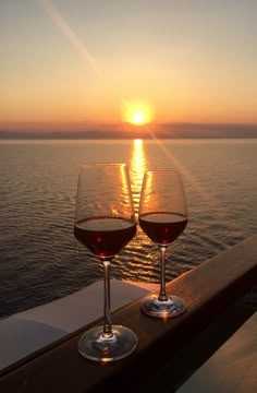 two glasses of wine sitting on top of a wooden table next to the ocean at sunset