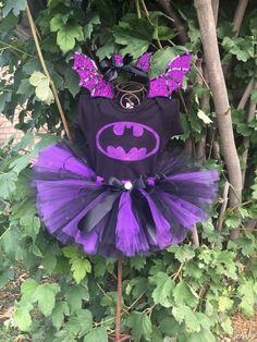 a purple and black batman tutu dress with ears on the top, in front of some green leaves