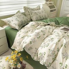 a bed with white sheets and green comforter next to a vase filled with yellow flowers