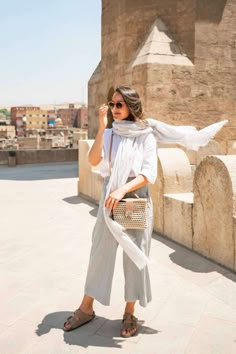 a woman in white shirt and grey pants holding a beige bag while standing next to an old building