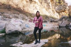 a woman in pink jacket standing on rock next to river with graffiti all over it