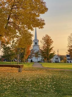 a park bench in front of a church with autumn leaves on the ground and trees surrounding it