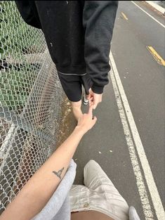 two people holding hands while standing on the side of a road near a chain link fence