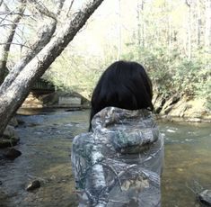 a woman standing in front of a river wearing a camo jacket and looking at the water