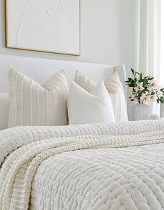 a white bed topped with pillows next to a vase filled with flowers and a painting on the wall