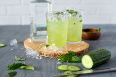 two glasses filled with cucumber and ice on top of a cutting board next to a bottle