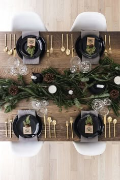 the table is set with black plates and silverware, pine cones, greenery