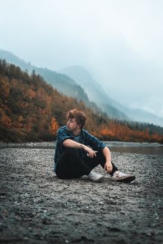 a man sitting on the ground next to a body of water with mountains in the background