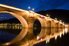 an old stone bridge is lit up at night with lights reflecting in the water below