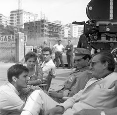 black and white photograph of people sitting on the sidewalk in front of a movie camera