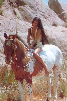 a woman riding on the back of a brown and white horse next to a rocky hillside