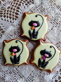 three decorated cookies sitting on top of a lace doily covered tablecloth with white and black cats