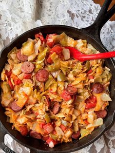 a skillet filled with cabbage and sausage on top of a lace table cloth next to a red spoon