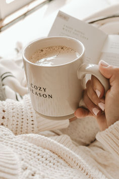 a woman is holding a cup of coffee and reading a book while laying in bed