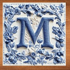 the letter m is made out of blue thread and has leaves on it, with an embroidered border