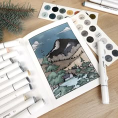 an open book with some markers and pens next to it on a wooden table, surrounded by other art supplies