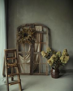 an old window with curtains and flowers in it next to a ladder, chair and vase