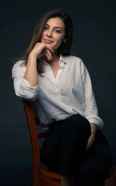 a woman sitting on top of a wooden chair in front of a dark background with her hand under her chin