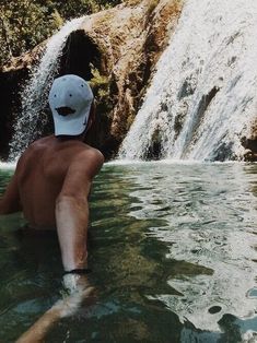 a man swimming in the water next to a waterfall with a hat on his head