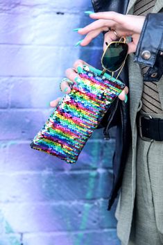 a woman wearing sunglasses and a jacket holding a colorful purse in front of a brick wall