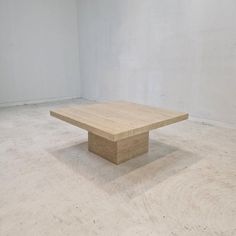 a square wooden table sitting on top of a cement floor in an empty room with white walls